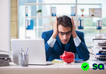 Hombre joven con traje azul mirando preocupado una alcancía vacía sobre su escritorio, rodeado de papeles y una computadora, simbolizando la necesidad urgente de un aumento de salario.