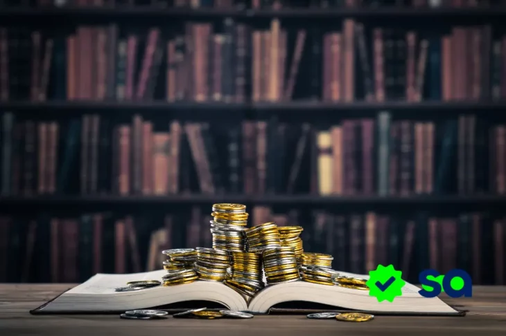 Pilas de monedas sobre un libro abierto con estantería de libros al fondo