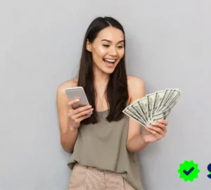 Mujer joven sonriendo mientras sostiene un abanico de billetes en una mano y un celular en la otra, representando la conexión entre dinero y emociones.