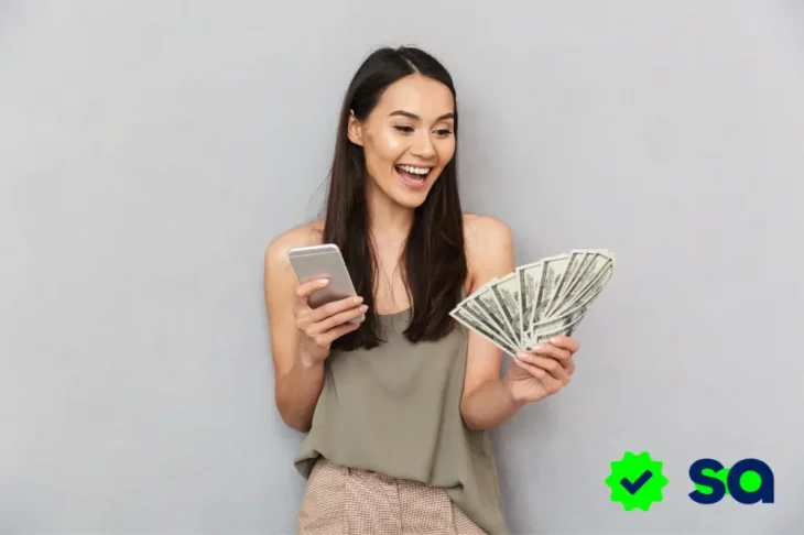 Mujer joven sonriendo mientras sostiene un abanico de billetes en una mano y un celular en la otra, representando la conexión entre dinero y emociones.
