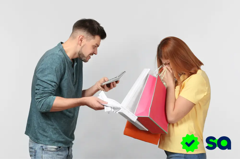 Hombre molesto revisando un recibo mientras confronta a una mujer con varias bolsas de compras, representando el acto de gastar para llenar vacíos emocionales.