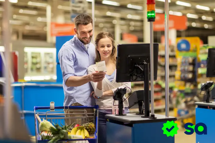 Pareja pagando en supermercado con carrito de compras lleno, utilizando vales de despensa.