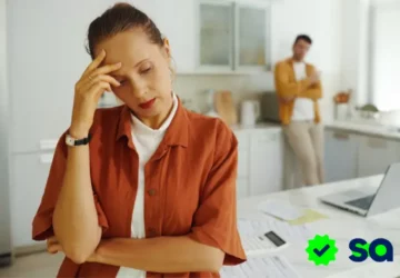 Mujer preocupada revisando documentos en la cocina mientras un hombre la observa al fondo, representando la duda sobre si te pueden embargar cuando no tienes bienes a tu nombre.
